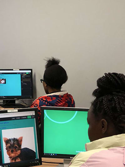 Adult stands in front of multiple young people at computers in classroom setting.